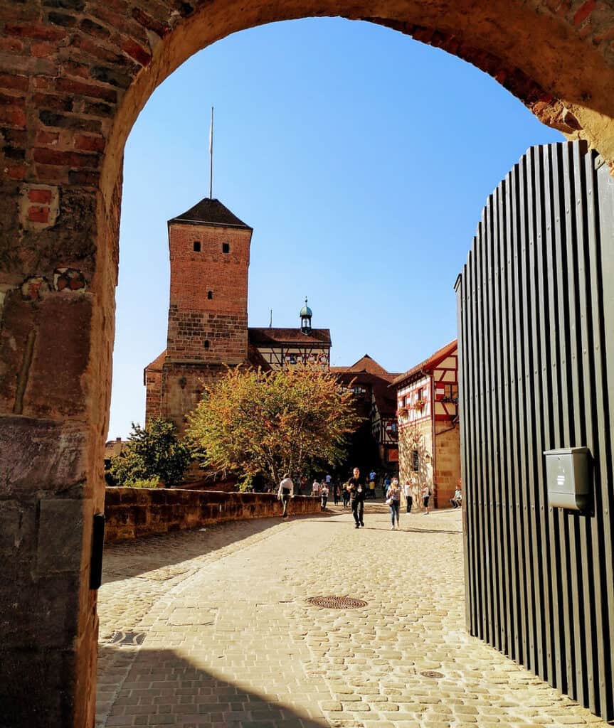 A large wooden gate is open in a stone arched wall. Behind the gate, a small tower with a pointed roof can be seen in the background.