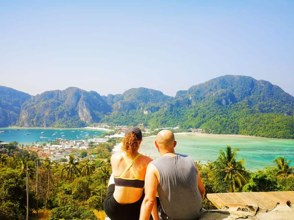 A man and woman sit on the ground facing out to a bright blue bay, framed by limestone mountains covered in green forest, in Koh Phi Phi, Thailand