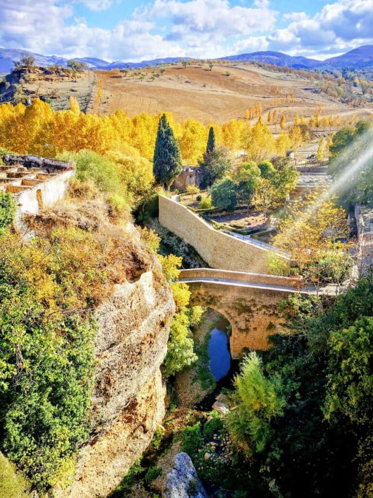 A small stone footbridge is surrounded by yellow and golden fields and trees in the town of Ronda, Spain