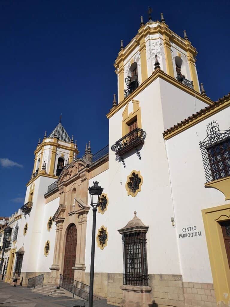 A white washed cathedral with yellow coloured detail around the edges and a modest clock tower, in the small town of Ronda, Spain