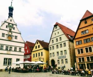 A row of orange, yellow and red houses surround a cobbled square. The houses have a terracotta pitched roofs and diners sit at café tables in the square, in Rothenberg ob de Tauber, Germany