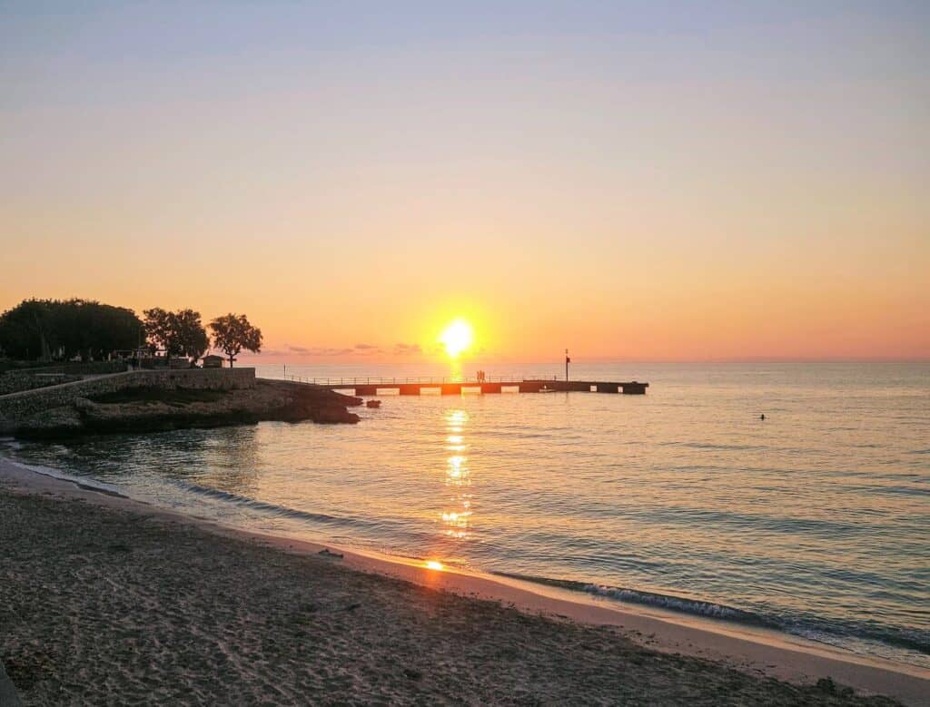 The sun rises above the calm sea. Silhouettes of two people can be seen walking along a small pier