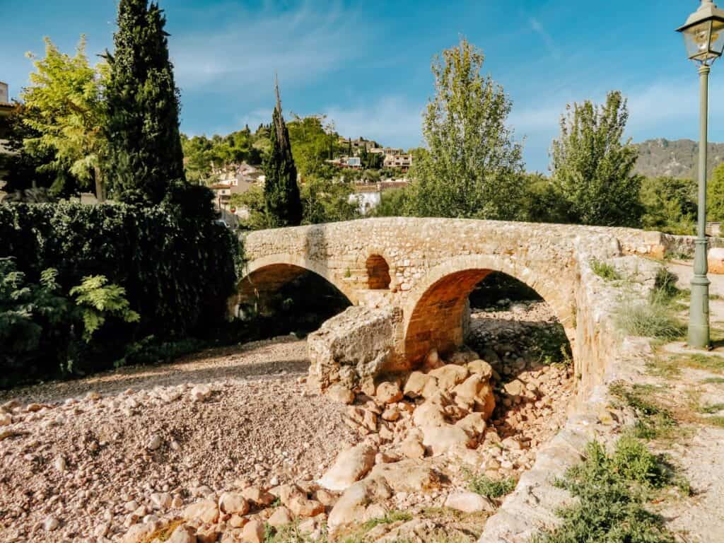 A small stone bridge arches over a dried-up stream, surrounded by green trees in Pollenca, Mallorca